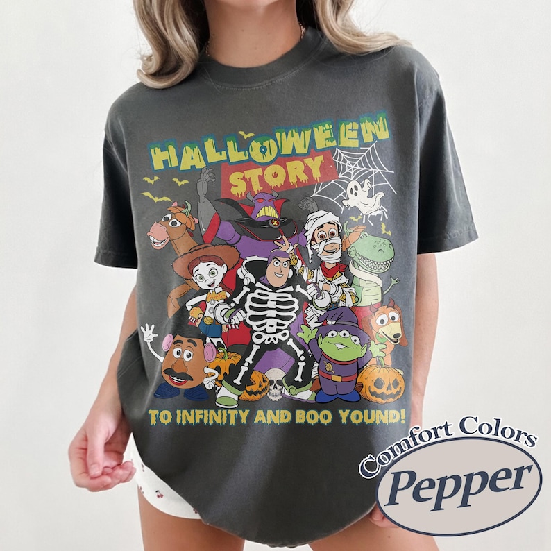 May include: Dark gray Comfort Colors t-shirt featuring a Halloween-themed graphic with the text "HALLOWEEN STORY" and "TO INFINITY AND BOO YOUND!" The graphic includes cartoon characters and pumpkins. The shirt has a crew neck.