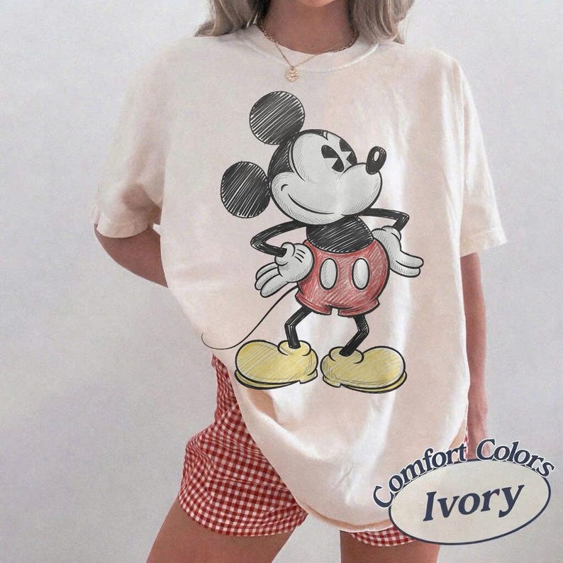 May include: Ivory-colored t-shirt featuring a sketch of Mickey Mouse in black, red, and yellow. The shirt has a relaxed fit and is paired with red and white checkered shorts. The text "Comfort Colors Ivory" is printed on the bottom right.