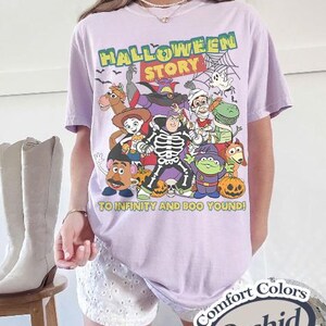 May include: A light purple t-shirt featuring a Halloween-themed graphic with various cartoon characters. The text "HALLOWEEN STORY" is at the top, with "TO INFINITY AND BOO YOND!" below. The shirt is labeled "Comfort Colors Orchid".