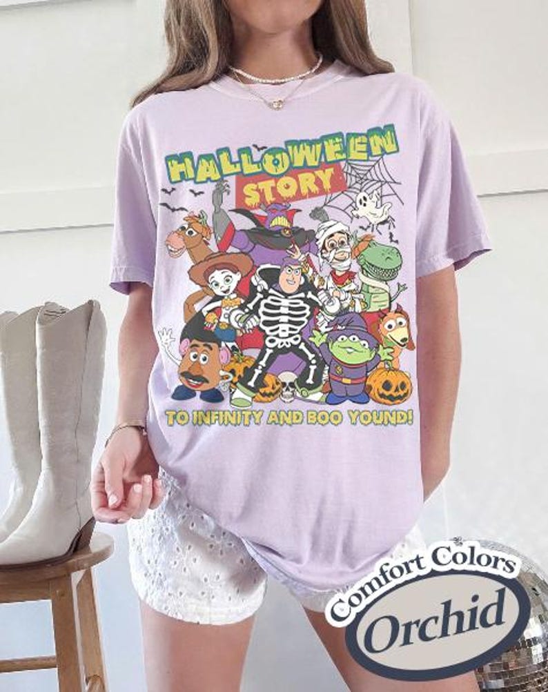 May include: A light purple t-shirt featuring a Halloween-themed graphic with various cartoon characters. The text "HALLOWEEN STORY" is at the top, with "TO INFINITY AND BOO YOND!" below. The shirt is labeled "Comfort Colors Orchid".