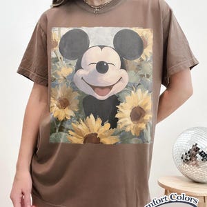 Puede incluir: Camiseta color espresso con Mickey Mouse sonriente rodeado de girasoles. El gr&aacute;fico est&aacute; centrado en la camiseta. La camiseta tiene mangas cortas y cuello redondo. Las palabras "Comfort Colors Espresso" est&aacute;n impresas en una etiqueta en la esquina inferior derecha.