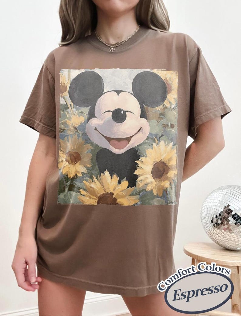Puede incluir: Camiseta color espresso con Mickey Mouse sonriente rodeado de girasoles. El gr&aacute;fico est&aacute; centrado en la camiseta. La camiseta tiene mangas cortas y cuello redondo. Las palabras "Comfort Colors Espresso" est&aacute;n impresas en una etiqueta en la esquina inferior derecha.