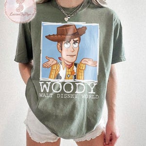 Puede incluir: Camiseta verde oliva con una imagen de dibujos animados de Woody de Toy Story con los brazos extendidos. La imagen est&aacute; sobre un fondo azul claro. La palabra "WOODY" est&aacute; impresa en blanco debajo, con "WALT DISNEY WORLD" debajo.