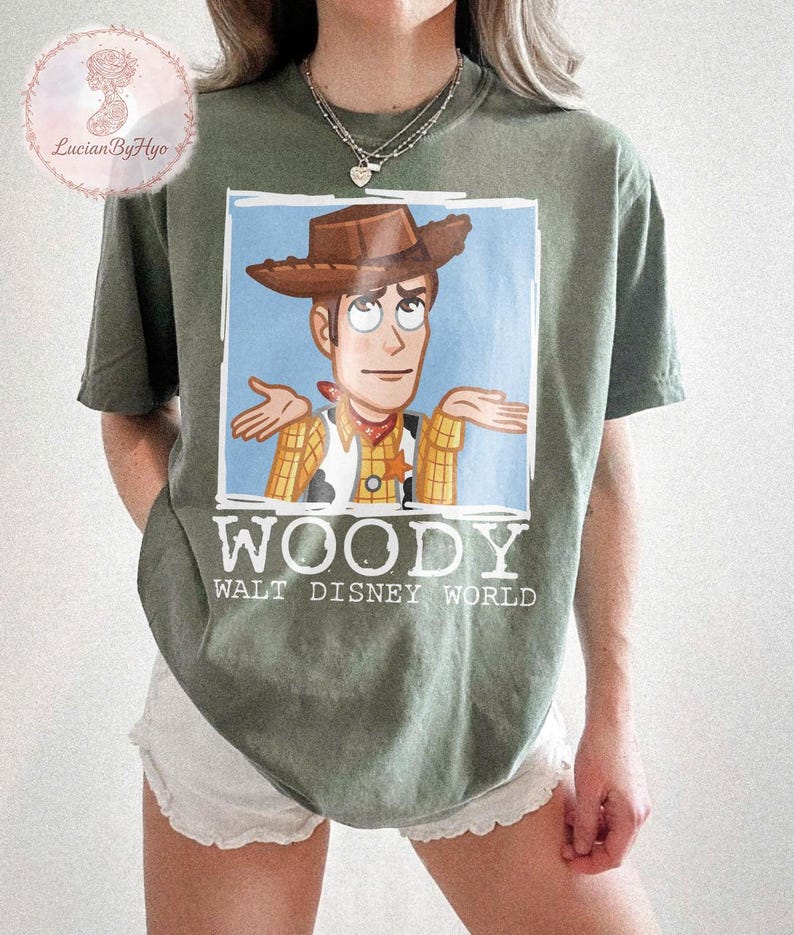 Puede incluir: Camiseta verde oliva con una imagen de dibujos animados de Woody de Toy Story con los brazos extendidos. La imagen est&aacute; sobre un fondo azul claro. La palabra "WOODY" est&aacute; impresa en blanco debajo, con "WALT DISNEY WORLD" debajo.