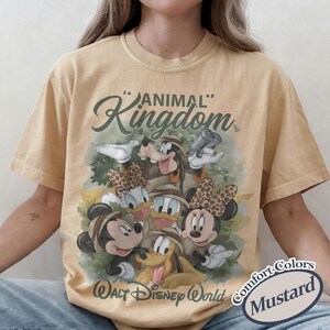 May include: Mustard-colored t-shirt featuring a graphic of Disney characters in safari attire. The design includes Mickey, Minnie, Donald, Daisy, Goofy, and Pluto, with "Animal Kingdom" and "Walt Disney World" text.