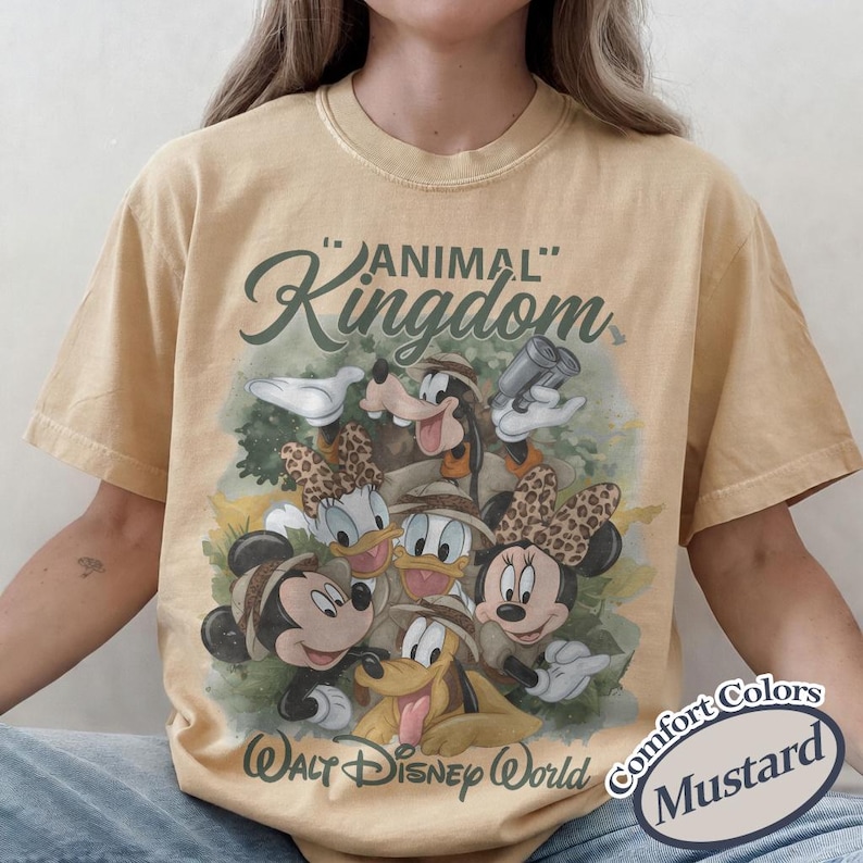 May include: Mustard-colored t-shirt featuring a graphic of Disney characters in safari attire. The design includes Mickey, Minnie, Donald, Daisy, Goofy, and Pluto, with "Animal Kingdom" and "Walt Disney World" text.