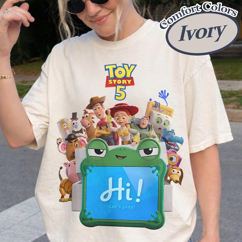 May include: An ivory t-shirt featuring a graphic print of Toy Story 5 characters including Woody, Jessie, and Buzz Lightyear. A green frog-shaped tablet frame is centered in the foreground with the text Hi! Let's play! printed on the screen.