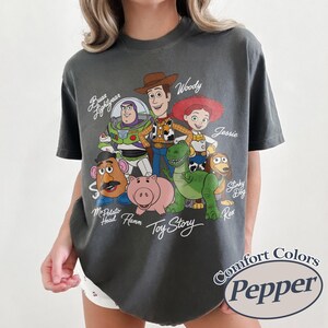 May include: Dark grey t-shirt with a graphic of Toy Story characters, including Woody, Buzz Lightyear, and Jessie. The shirt has the words "Toy Story" in a script font. The shirt also has the words "Comfort Colors Pepper" in a circle.