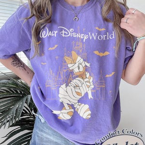 以下が含まれることがあります： 紫色のComfort Colors Tシャツ。お城の前でミイラ姿のドナルドダックのグラフィック、テキスト「Walt Disney World」入り。コウモリと「Violet」の文字も含まれています。