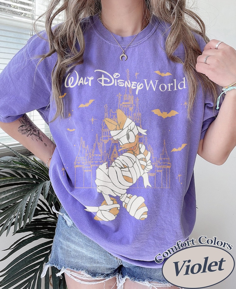 以下が含まれることがあります： 紫色のComfort Colors Tシャツ。お城の前でミイラ姿のドナルドダックのグラフィック、テキスト「Walt Disney World」入り。コウモリと「Violet」の文字も含まれています。