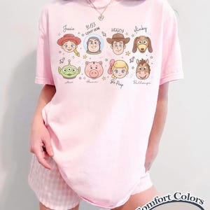 May include: A light pink t-shirt featuring cartoon character faces from the Toy Story movie. The shirt includes the names Jessie, Buzz Lightyear, Woody, Slinky, Alien, Hamm, Bo Peep, and Bullseye. The text "Comfort Colors Blossom" is printed on the bottom right.