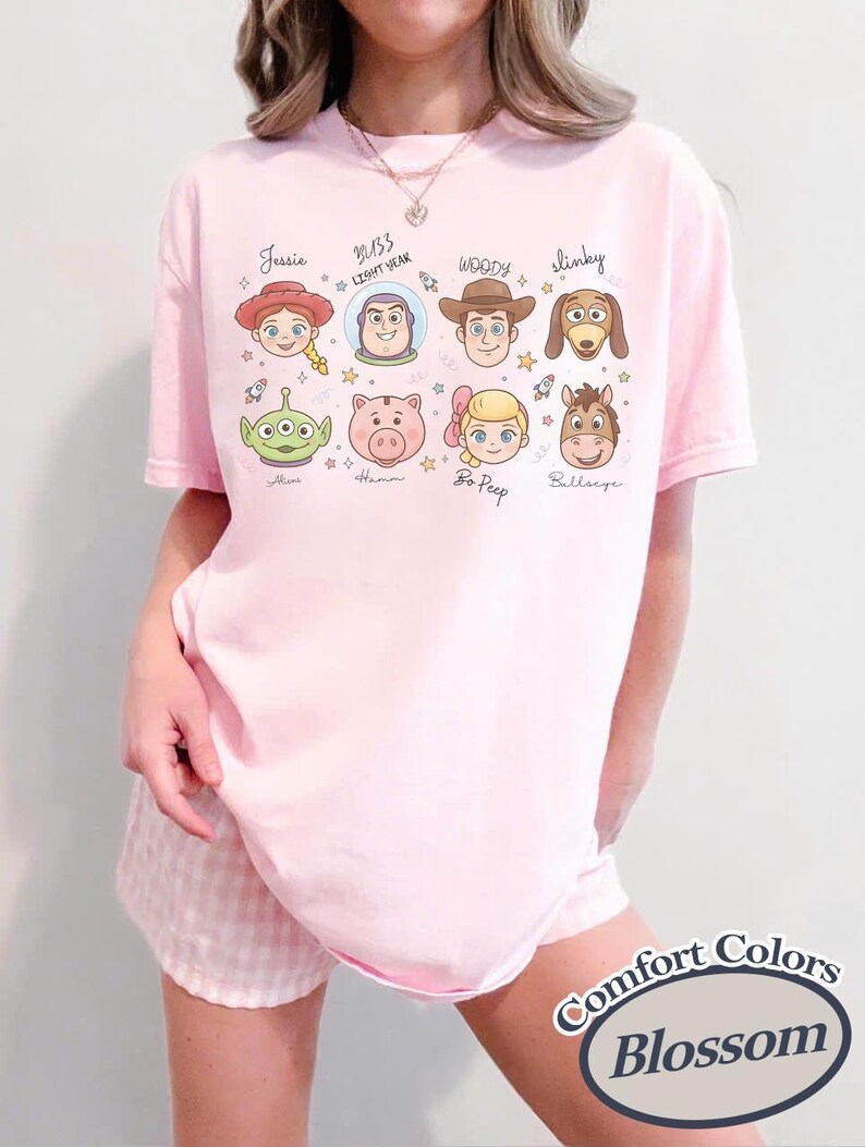 May include: A light pink t-shirt featuring cartoon character faces from the Toy Story movie. The shirt includes the names Jessie, Buzz Lightyear, Woody, Slinky, Alien, Hamm, Bo Peep, and Bullseye. The text "Comfort Colors Blossom" is printed on the bottom right.