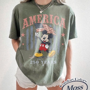 Pu&ograve; includere: T-shirt verde oliva con Topolino che tiene una bandiera americana. La parola "AMERICA" &egrave; sopra, con "SINCE 1776" e "250 YEARS" sotto. La maglietta ha un aspetto vintage.