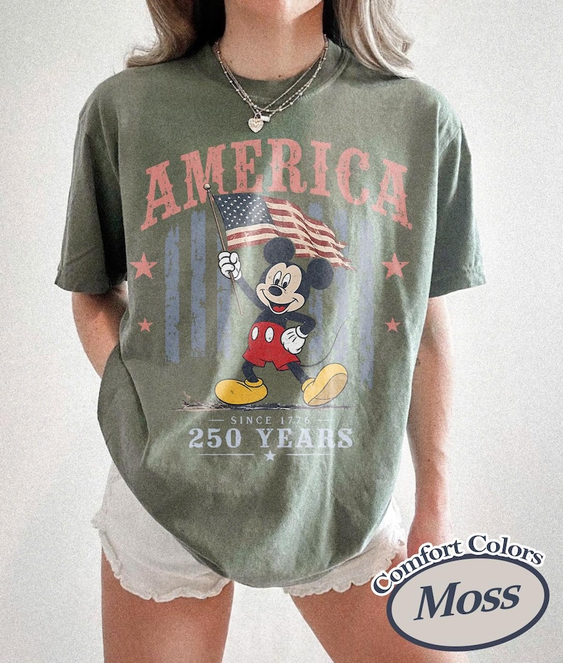 Pu&ograve; includere: T-shirt verde oliva con Topolino che tiene una bandiera americana. La parola "AMERICA" &egrave; sopra, con "SINCE 1776" e "250 YEARS" sotto. La maglietta ha un aspetto vintage.