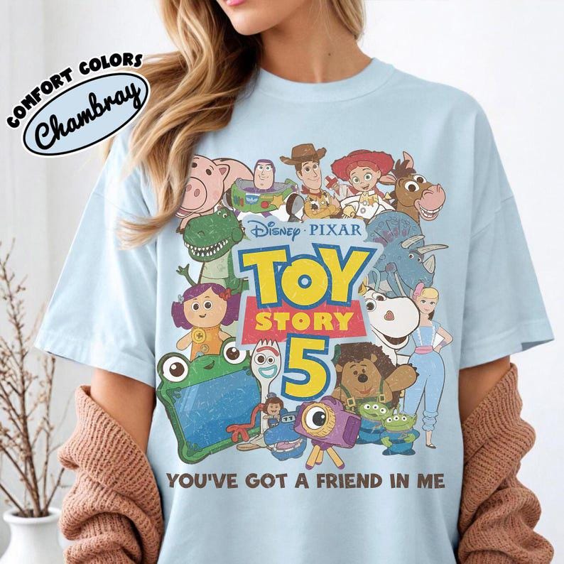 May include: A light blue t-shirt featuring a graphic print of various Toy Story characters, the Disney Pixar Toy Story 5 logo, and the text You've Got A Friend In Me. The shirt includes a circular label in the corner that reads Comfort Colors Chambray.