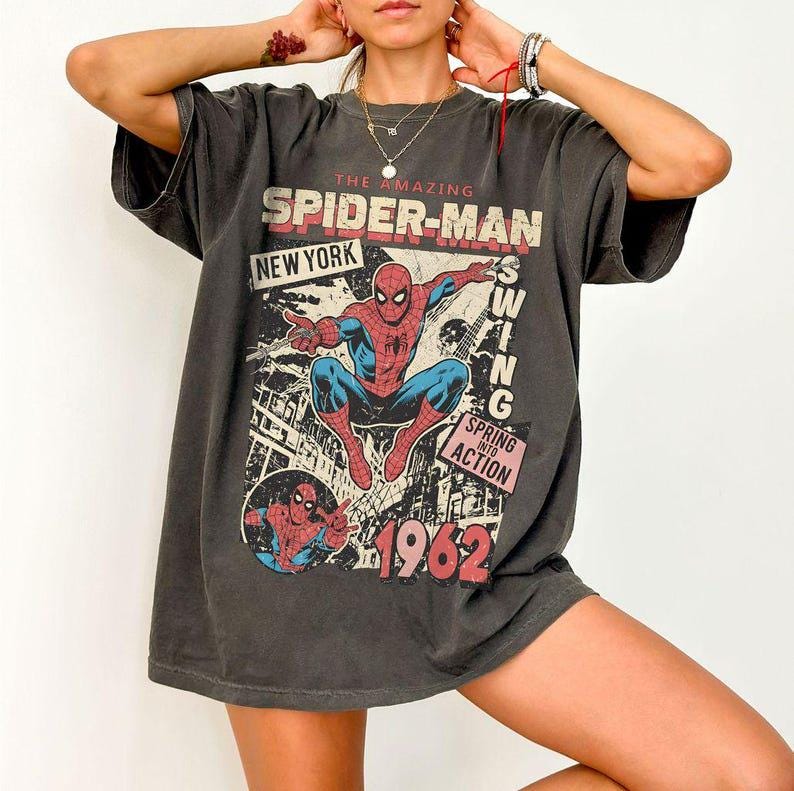 May include: A charcoal gray short-sleeve t-shirt featuring a vintage-style graphic of Spider-Man. The print includes the text THE AMAZING SPIDER-MAN, NEW YORK, SWING, SPRING INTO ACTION, and the year 1962 in red and white lettering over a city scene.