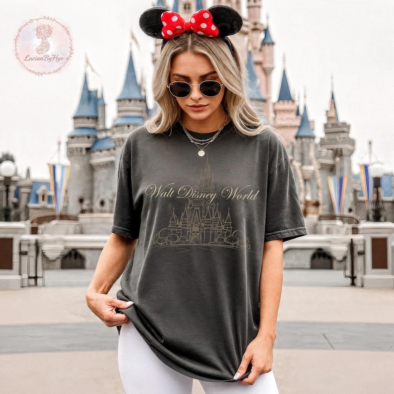 May include: Dark gray t-shirt with the text "Walt Disney World" and a castle graphic. The shirt is worn with black sunglasses and a Minnie Mouse ear headband with a red polka dot bow. The background features a castle.