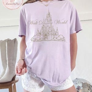 May include: A light purple t-shirt featuring a gold-colored illustration of Cinderella's castle with the text "Walt Disney World". The shirt is a relaxed fit and has short sleeves. The shirt is paired with white shorts.