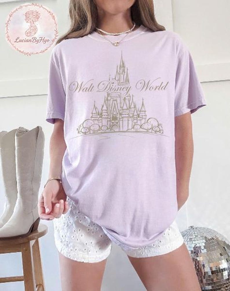 May include: A light purple t-shirt featuring a gold-colored illustration of Cinderella's castle with the text "Walt Disney World". The shirt is a relaxed fit and has short sleeves. The shirt is paired with white shorts.