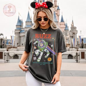 May include: Dark gray t-shirt featuring a graphic of Buzz Lightyear with the word "BUZZ" in red. The shirt also includes the text "YOU'VE GOT A FRIEND IN ME" and "Est. 1997". The design is set against a vintage-style frame.