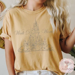 May include: Mustard yellow t-shirt featuring a gray illustration of a castle and the words "Walt Disney World" in a script font. The shirt has short sleeves and a relaxed fit.