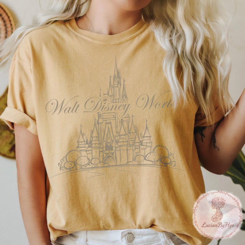 May include: Mustard yellow t-shirt featuring a gray illustration of a castle and the words "Walt Disney World" in a script font. The shirt has short sleeves and a relaxed fit.