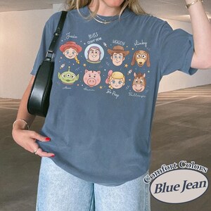 May include: Blue jean colored t-shirt featuring cartoon character faces from a popular animated movie. The shirt includes the names of the characters: Jessie, Buzz Lightyear, Woody, Slinky, Alien, Hamm, Bo Peep, and Bullseye.