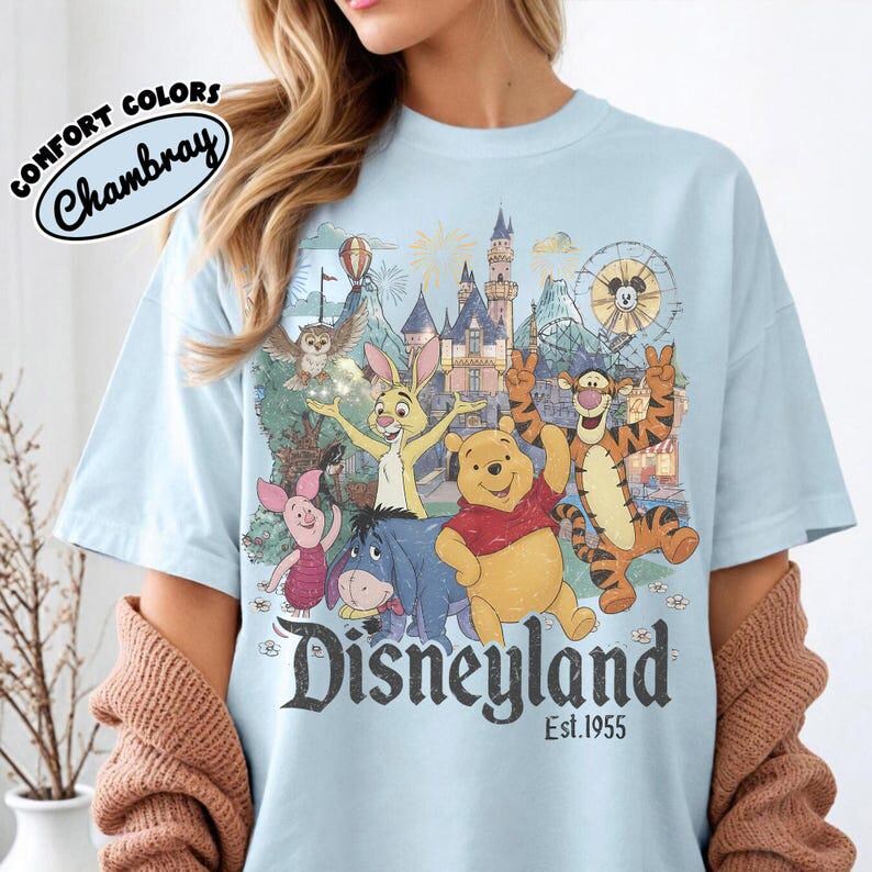 May include: A light blue t-shirt featuring a graphic print of Winnie the Pooh, Tigger, Piglet, Eeyore, and Rabbit in front of a castle. The text reads Disneyland Est. 1955. A circular logo in the top left corner says Comfort Colors Chambray.