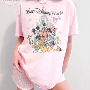 May include: Pink t-shirt featuring the text "Walt Disney World" above a colorful graphic of Mickey Mouse, Minnie Mouse, Goofy, Donald Duck, Daisy Duck, and Chip and Dale. The shirt is a light pink color.