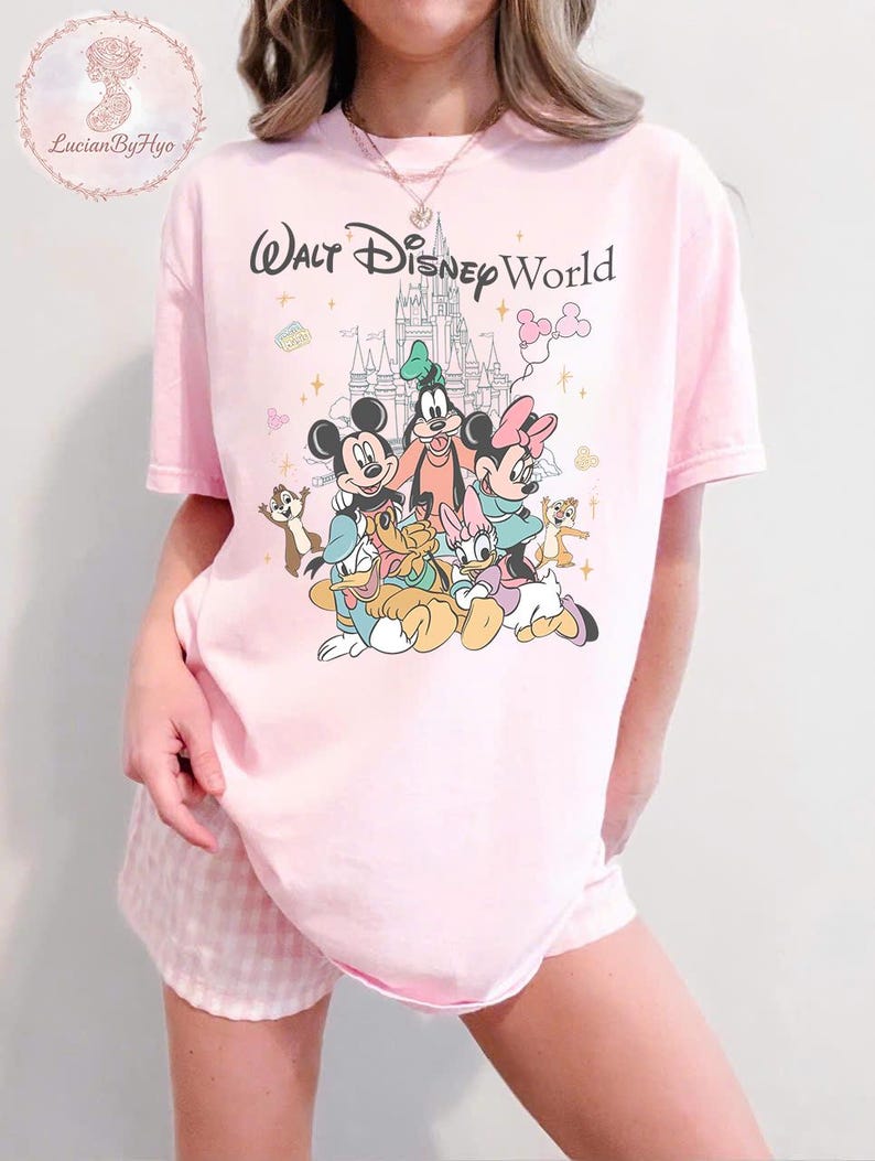 May include: Pink t-shirt featuring the text "Walt Disney World" above a colorful graphic of Mickey Mouse, Minnie Mouse, Goofy, Donald Duck, Daisy Duck, and Chip and Dale. The shirt is a light pink color.