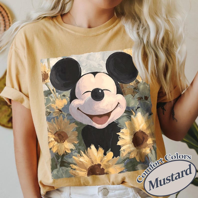 Puede incluir: Camiseta de color mostaza con un gr&aacute;fico de Mickey Mouse sonriente rodeado de girasoles. El gr&aacute;fico es una ilustraci&oacute;n de estilo pintura. La camiseta tiene las mangas enrolladas y las palabras "Comfort Colors" y "Mustard" est&aacute;n impresas en la parte inferior derecha.