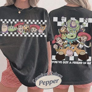 May include: Dark gray t-shirt featuring characters from the animated film Toy Story. The front of the shirt displays a row of character faces above a checkered pattern. The back of the shirt has a larger graphic of the characters with the text "YOU'VE GOT A FRIEND IN ME".