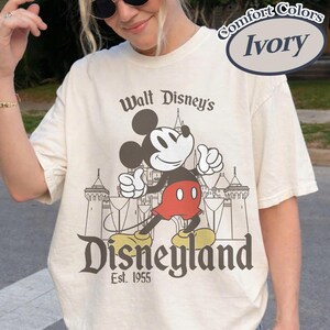 Puede incluir: Camiseta de color marfil con un gr&aacute;fico de Mickey Mouse haciendo una se&ntilde;al de aprobaci&oacute;n frente a un castillo, con las palabras "Walt Disney's Disneyland Est. 1955". La camiseta es de la marca Comfort Colors.