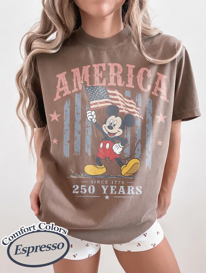 Pu&ograve; includere: T-shirt marrone con la scritta "AMERICA" in rosa, una grafica di Mickey Mouse con la bandiera americana e il testo "SINCE 1776" e "250 YEARS". La maglietta &egrave; color Espresso.