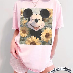 Puede incluir: Camiseta rosa claro con un dise&ntilde;o de Mickey Mouse sonriente rodeado de girasoles. La camiseta es un modelo Comfort Colors Blossom. La persona tambi&eacute;n lleva un pantal&oacute;n corto rosa a cuadros.