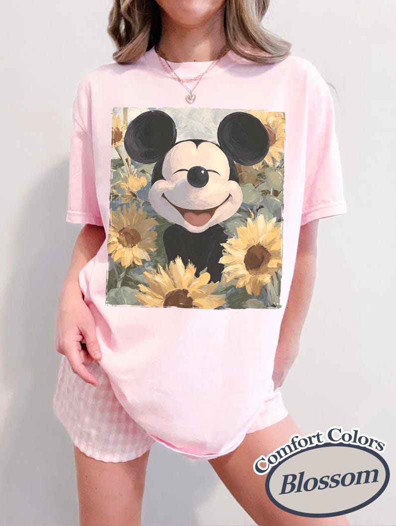 Puede incluir: Camiseta rosa claro con un dise&ntilde;o de Mickey Mouse sonriente rodeado de girasoles. La camiseta es un modelo Comfort Colors Blossom. La persona tambi&eacute;n lleva un pantal&oacute;n corto rosa a cuadros.