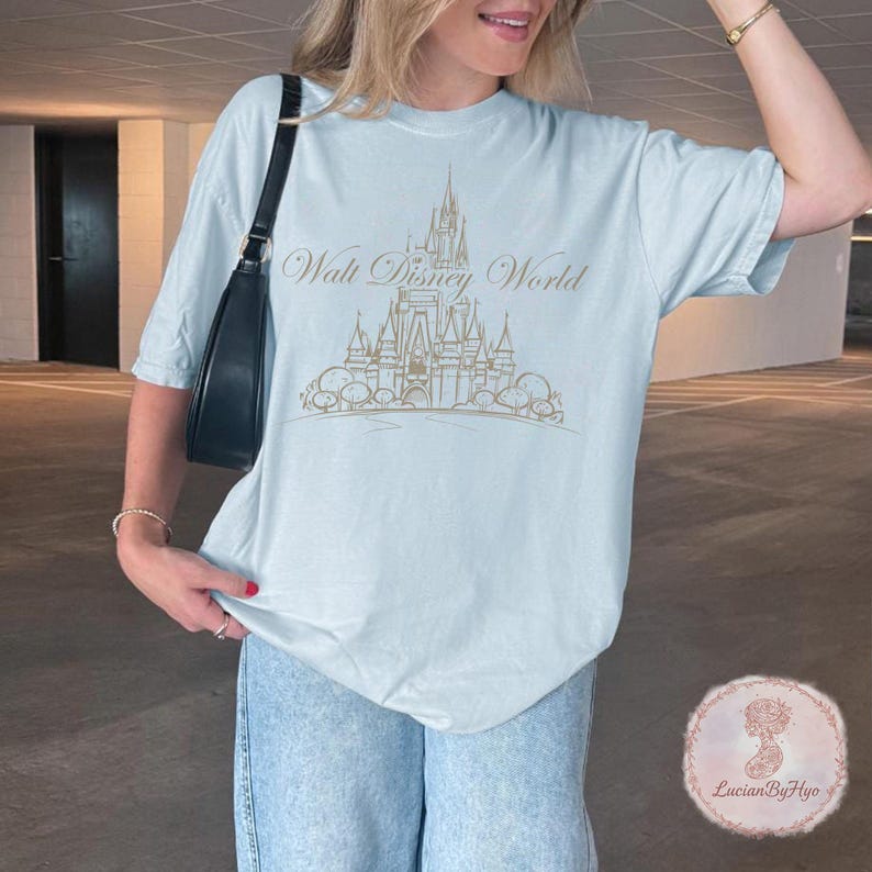 May include: Light blue t-shirt with a gold illustration of a castle and the words "Walt Disney World". The shirt is a soft, comfortable material and has short sleeves. The shirt is a light blue color.