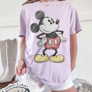 May include: A light purple t-shirt featuring a classic Mickey Mouse illustration. The cartoon character is depicted in black, red, yellow, and white. The shirt is labeled "Comfort Colors Orchid".