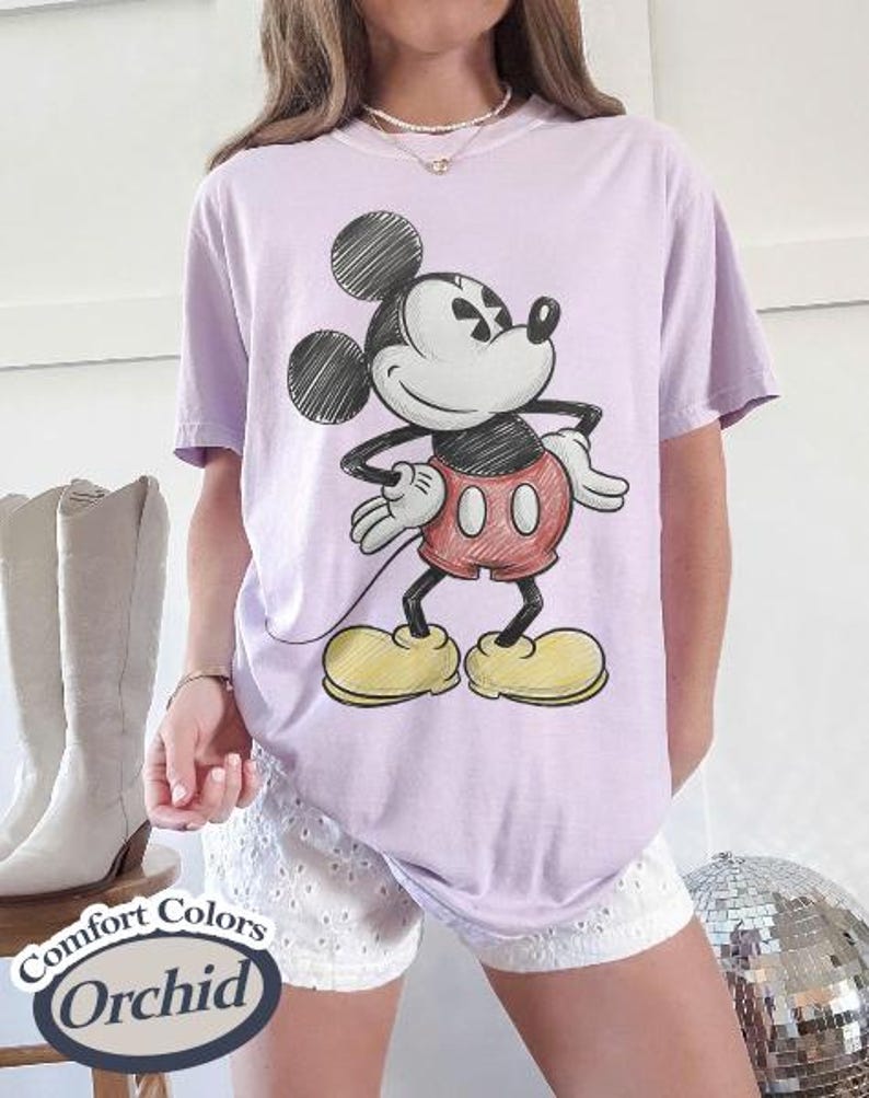 May include: A light purple t-shirt featuring a classic Mickey Mouse illustration. The cartoon character is depicted in black, red, yellow, and white. The shirt is labeled "Comfort Colors Orchid".