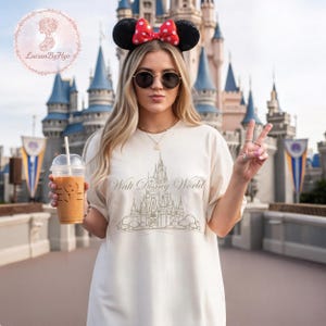 May include: Cream-colored t-shirt with a gold-colored graphic of a castle and the words "Walt Disney World." The person is wearing Minnie Mouse ears with a red bow and sunglasses. They are holding a beverage and making a peace sign.