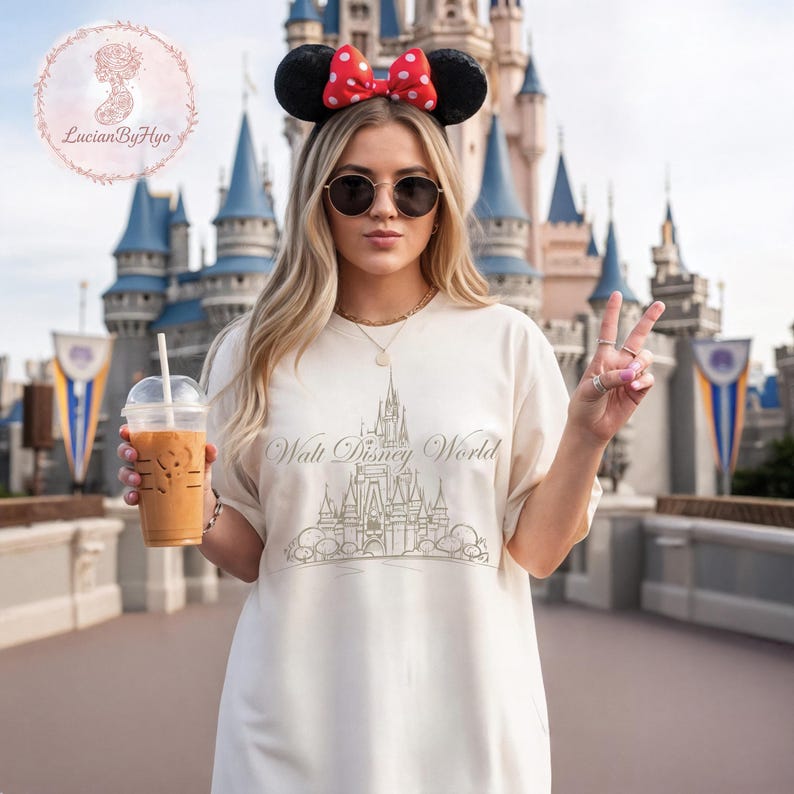 May include: Cream-colored t-shirt with a gold-colored graphic of a castle and the words "Walt Disney World." The person is wearing Minnie Mouse ears with a red bow and sunglasses. They are holding a beverage and making a peace sign.