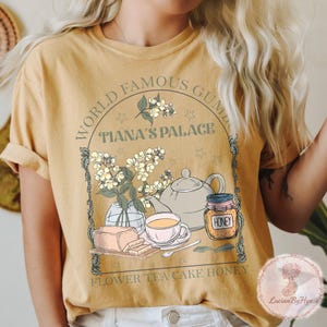 May include: Mustard yellow t-shirt with a graphic design featuring a teapot, teacup, honey jar, and flowers. The text reads "World Famous Gumbo Tiana's Palace" and "Flower Tea Cake Honey."