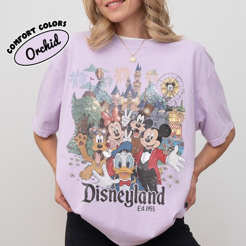 Peut inclure: Un t-shirt violet clair avec une impression graphique de Mickey, Minnie, Donald, Dingo et Pluto devant un ch&acirc;teau. Le texte indique COMFORT COLORS Orchid, Disneyland et Est. 1955. Le motif comprend des feux d'artifice et une grande roue.