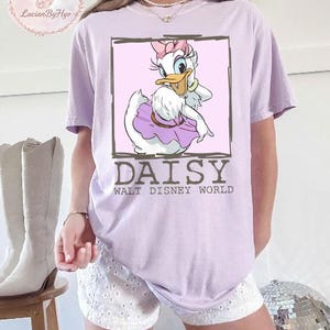 Puede incluir: Camiseta lila claro con un dise&ntilde;o de Daisy Duck de Walt Disney World. Daisy aparece en un marco rosa, con un lazo rosa y una falda morada. El texto "DAISY WALT DISNEY WORLD" est&aacute; debajo.