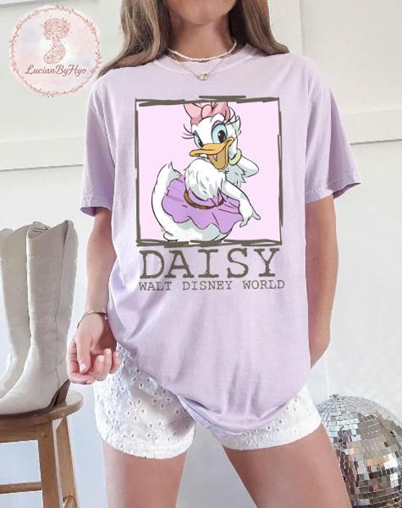 Puede incluir: Camiseta lila claro con un dise&ntilde;o de Daisy Duck de Walt Disney World. Daisy aparece en un marco rosa, con un lazo rosa y una falda morada. El texto "DAISY WALT DISNEY WORLD" est&aacute; debajo.