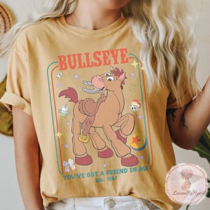May include: Mustard yellow t-shirt featuring a cartoon image of Bullseye, a horse character. The design includes the word "BULLSEYE" above the horse and the phrase "YOU'VE GOT A FRIEND IN ME" below. The shirt is a vintage style.