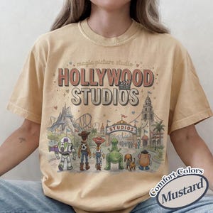 May include: Mustard-colored t-shirt featuring the text "HOLLYWOOD STUDIOS" with a graphic of theme park characters and attractions. The shirt also has the text "magic picture studio" and "Comfort Colors".