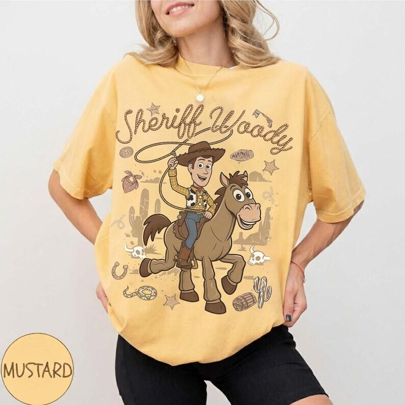 May include: A mustard yellow t-shirt featuring a graphic print of Sheriff Woody riding a horse. The design includes the text "Sheriff Woody" in a cursive font, surrounded by desert-themed illustrations like cacti, skulls, and lassos.