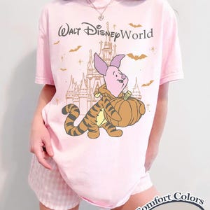 以下が含まれることがあります： ピンク色のTシャツ。プーさんのピグレットとティガーがカボチャを運び、お城を背景にしたグラフィック。グラフィックの上には「Walt Disney World」の文字。シャツには「Comfort Colors Blossom」と記載。