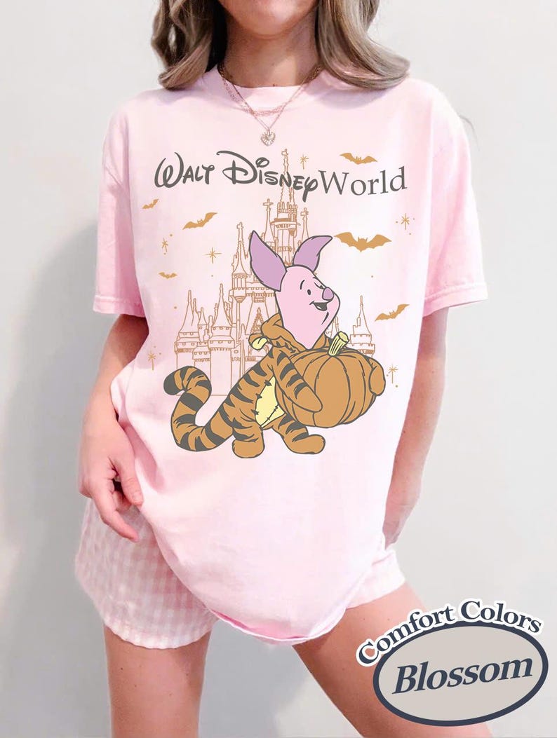 以下が含まれることがあります： ピンク色のTシャツ。プーさんのピグレットとティガーがカボチャを運び、お城を背景にしたグラフィック。グラフィックの上には「Walt Disney World」の文字。シャツには「Comfort Colors Blossom」と記載。